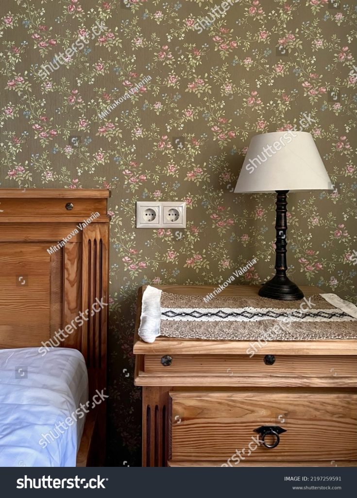 A cosy room for residents in the hotel. Green floral wallpaper, wooden bedside table and bed, night light and fresh white bed linen.